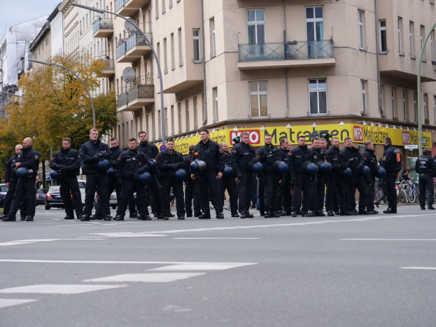 Gruppe von Polizisten in schwarzen Uniformen mit blauen Helmen, die auf einer Straße mit Laternen, Bäumen und Glasfenstern unter einem klaren blauen Himmel stehen.