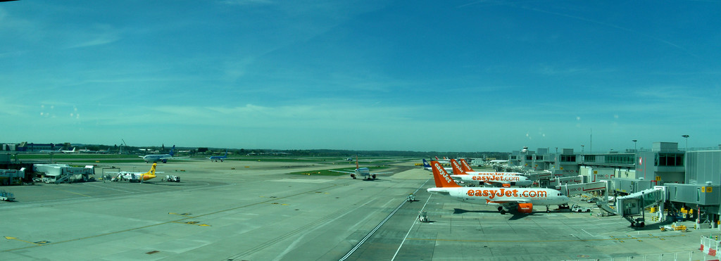 Eine Flughafen-Szene mit Flugzeugen, Gebäuden, Polen, Fahrzeugen, Bäumen, Himmel und einer Straße sowie einem Zaun im Vordergrund.