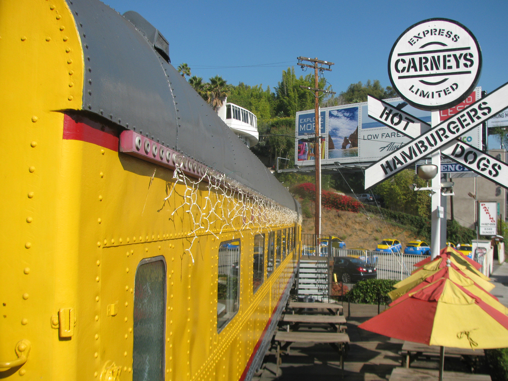 Ein mobiles Kantinengebäude mit Bänken und Stufen, umgeben von Schirmen, Pflanzen, einem Geländer, Fahrzeugen, einem Pfosten, Schildern, einem Gebäude, Bäumen und verschiedenen Gegenständen unter einem Himmel.