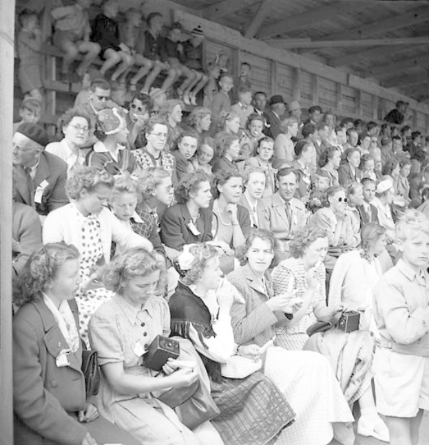 Schwarzes und weißes Foto einer Menge, die in einem Stadion sitzt, mit einer hölzernen Wand und einem Dach im Hintergrund, einige halten Kameras während eines Sportereignisses.