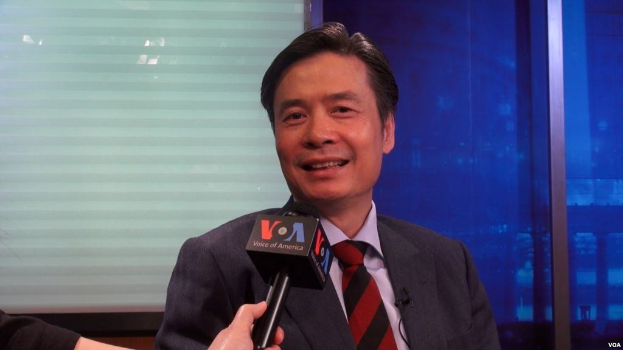 Ein Mann in Anzug und Krawatte hält ein Mikrofon, mit der Hand eines Reporters auf der linken Seite, vor einem Fenster und einer Wand.