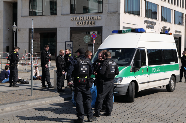 Polizeibeamte vor einem Starbucks-Café mit einem Van auf der rechten Seite und Menschen auf der linken Seite, vor einem Gebäude, einem Schild, einem Laternenpfahl und einem Zaun.