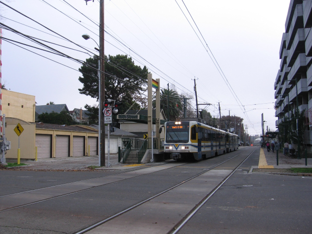 Straßenbahn fährt durch eine Stadtstraße mit hohen Gebäuden, Strommasten, Fußgängern, Bäumen und einem klaren blauen Himmel.