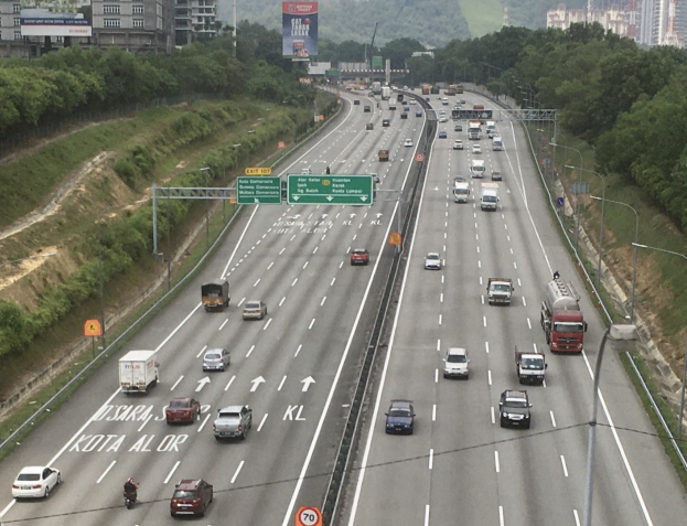 Eine belebte Autobahn mit hohen Gebäuden, Verkehrszeichen, Laternen und Bäumen an den Seiten, vor einem Hügel- und einem klaren blauen Himmel.