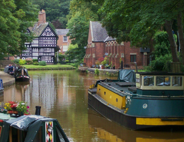 Kanal mit mehreren Booten, umgeben von grünem Gras, Pflanzen und Bäumen, mit Fenstern aufweisenden Gebäuden im Hintergrund.