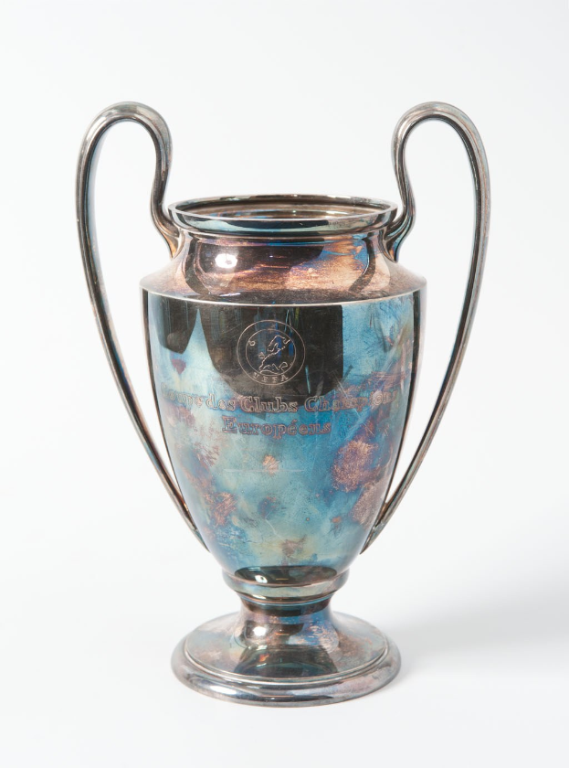 Der UEFA-Champions-League-Pokal, ein stahlerner Becher mit zwei Griffen und einem blauen und silbernen Farbschema, mit der Inschrift "UEFA Champions League" auf einem weißen Hintergrund.