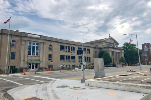 Ein großes Gebäude mit zahlreichen Fenstern, identifiziert als das New York State Penitentiary, umgeben von Straßenmasten, Straßenlaternen, Verkehrspfosten, Verkehrskegeln, Kraftfahrzeugen, Büschen, Bäumen und einem bewölkten Himmel.