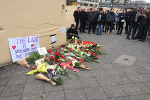 Gruppe von Menschen vor einem Zelt mit einer Tafel, auf der "Das Licht ist stärker als die Dunkelheit" steht, umgeben von Blumen, Kerzen und anderen Huldigungen, in einem öffentlichen Raum mit Gebäuden und Bäumen im Hintergrund.