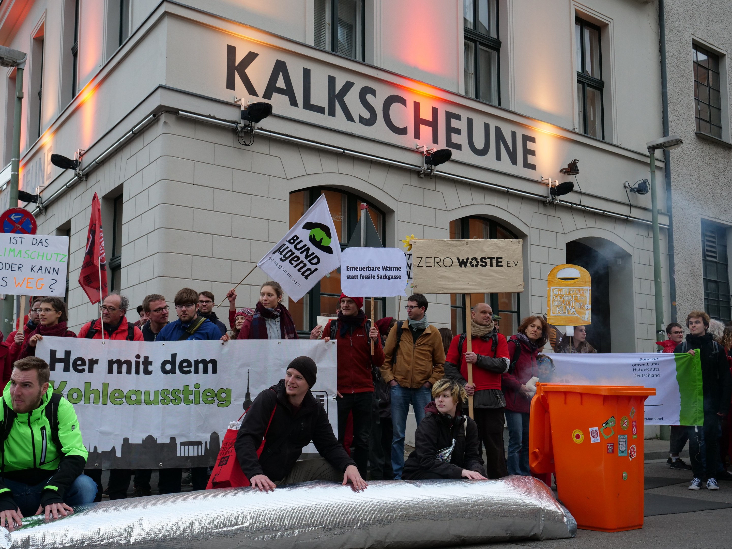 Eine Gruppe von Menschen mit Schildern und Plakaten vor einem Gebäude während einer Demonstration in Deutschland, mit zwei Personen auf einem Objekt im Vordergrund und einem Müllcontainer rechts.