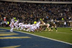 Ein Fußballspiel im Gange mit Spielern auf dem Rasen in Sportkleidung und Helmen, aus der Perspektive der Tribüne.