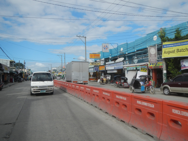 Eine belebte Stadtstraße mit mehreren Fahrzeugen, einer Mittelinsel, Gebäuden mit Beschilderung, Strommasten mit Drähten, Bäumen und einem bewölkten Himmel.