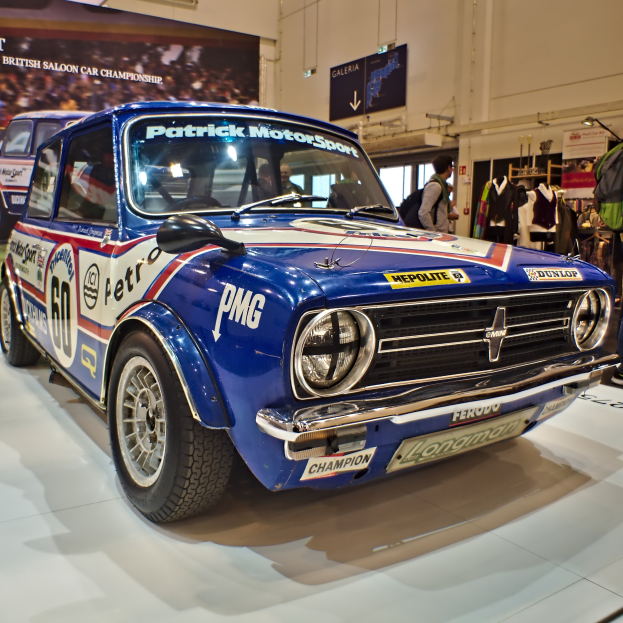 Kleiner blauer und weißer Mini Cooper S Rallye-Wagen in einem Museum ausgestellt, umgeben von Menschen mit Taschen, mit einem Banner und Beleuchtung im Hintergrund.