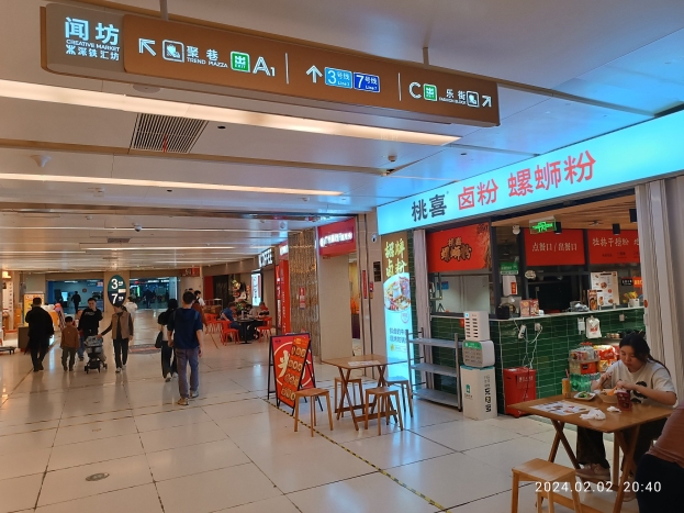 Menschen gehen durch ein Einkaufszentrum mit Tischen, auf denen Gegenstände ausgelegt sind, Texttafeln an den Wänden, Deckenlampen und einem zentralen kreativen Markt mit Essensauslagen.