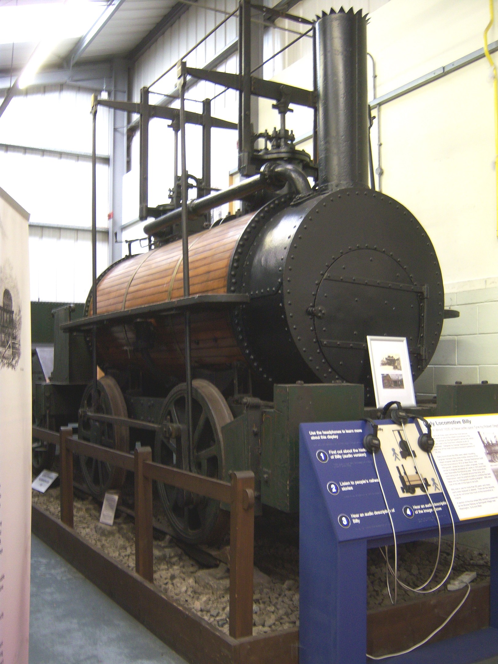 Alte Dampflokomotive in einem Museum mit Informationsschildern, einem Zaun, Steinen, einer Fahne, einer Wand mit Rohren und Deckenlampen.