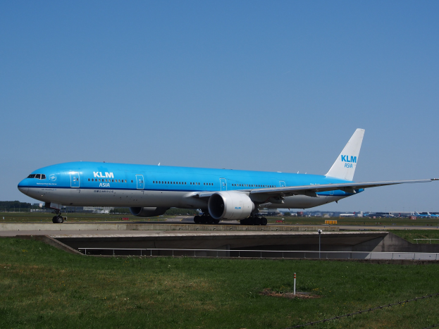 KLM Boeing 777-300ER auf der Rollbahn am Amsterdam Airport mit umliegender Wiese, Zaun, Pfählen, Gebäuden, Bäumen und einem klaren blauen Himmel.