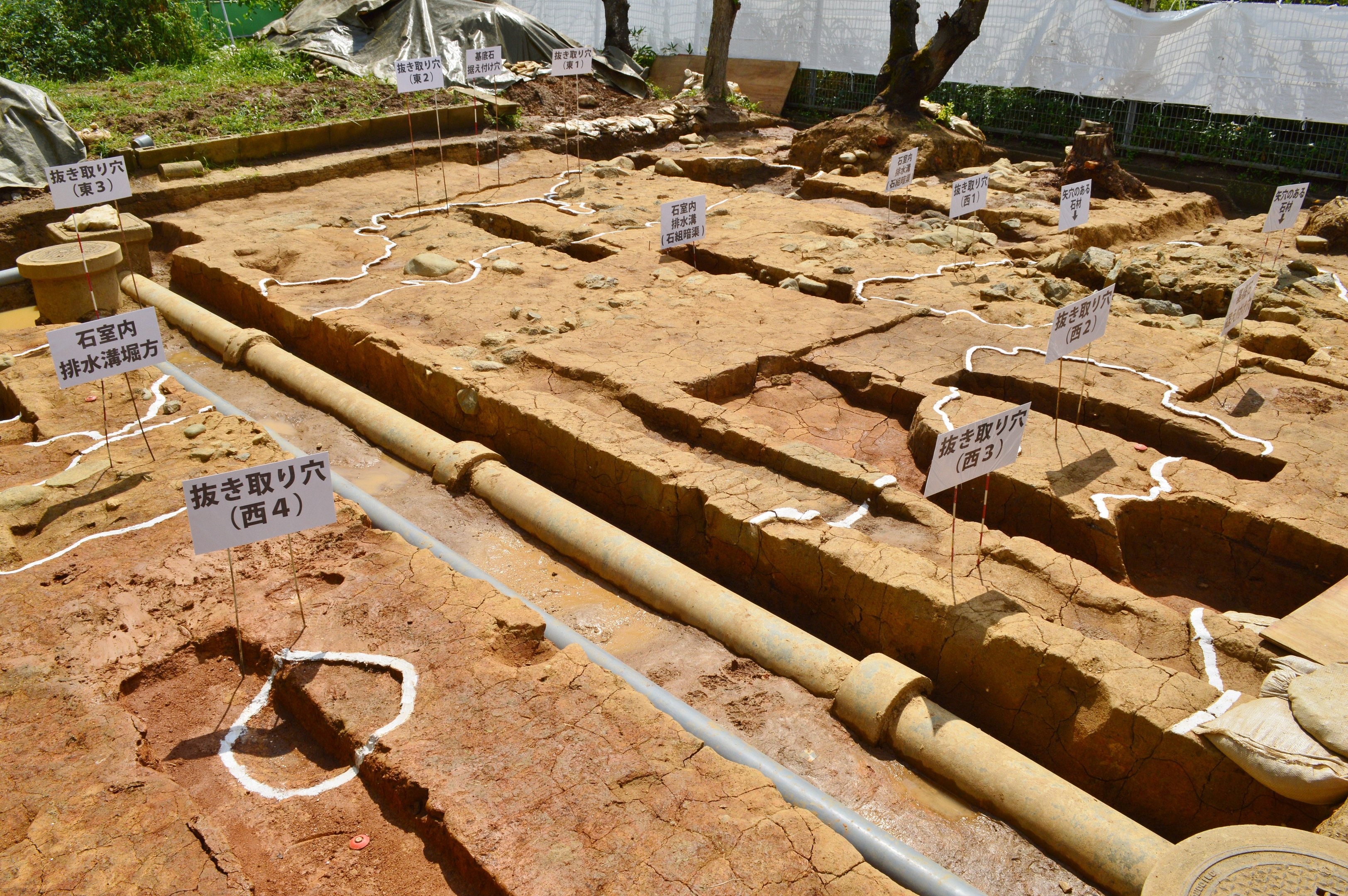 Großer Aushub mit freigelegter Erde, umgeben von Vegetation, einem Zaun, einer Abdeckung, einer Wand, Texttafeln, Rohren und verstreuten Gegenständen.