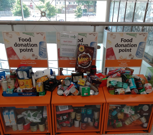 Orange Schränke mit Lebensmittelspenden, darunter Flaschen, Dosen und Kartons, mit Informationsschildern, durch Glasfenster betrachtet, die Fahrzeuge, Gebäude, Bäume und Himmel zeigen.