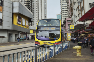 Ein gelber Doppeldeckerbus fährt eine Straße mit hohen Gebäuden entlang, mit Passanten auf dem rechten Gehweg, einem Hydranten und einem Geländer, unter einem klaren blauen Himmel.