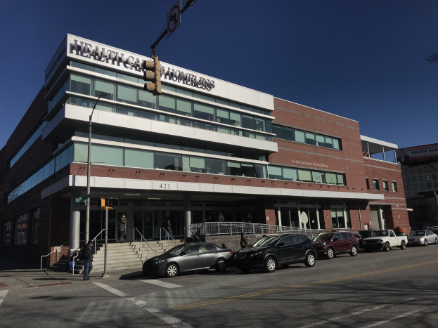 Großes Gebäude mit der Aufschrift "New York City Health Care Homeless Center" mit parkenden Autos davor, umgeben von Straßenmöbeln und Fußgängern unter einem klaren blauen Himmel.