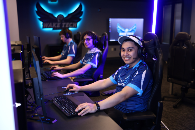 Eine Gruppe von Menschen in Headsets sitzt an Computern und lächelt, mit Monitoren, Tastaturen und Mäusen auf dem Schreibtisch, einem Logo an der Wand hinter ihnen und Stühlen rechts.