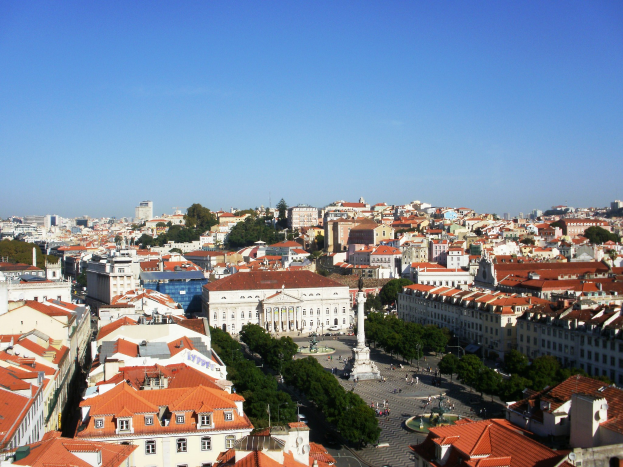 Ausblick auf Lissabon von einem Hügel mit Gebäuden mit Fenstern, Bäumen, einer Statue auf einem Sockel und einigen Menschen auf der Straße, mit dem Himmel im Hintergrund.