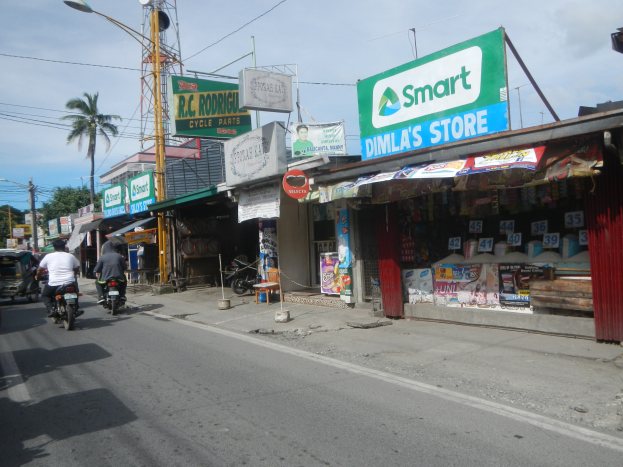 Eine belebte Straße mit Motorradfahrern, Autos, Gebäuden, Straßenschildern, Strommasten, Kabeln, Lichtern, Bäumen und einem bewölkten Himmel.