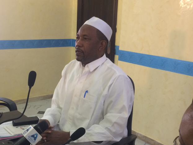 Ein Mann in einem weißen Gewand und einer Mütze sitzt an einem Tisch mit Mikrofonen, spricht, während Papiere und Gegenstände auf dem Tisch sichtbar sind, mit einer anderen Person mit Brille rechts von ihm, in einem Raum mit einer Wand und einer Tür im Hintergrund.