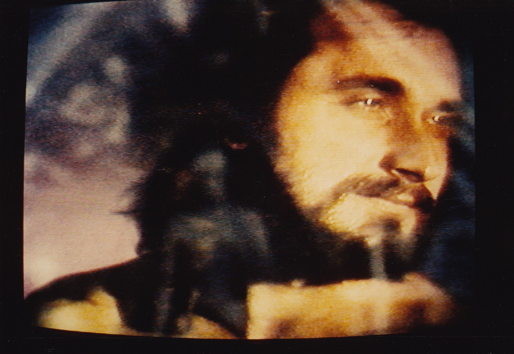 Ein Mann mit langem dunklem Bart und Haar, der ein weißes Hemd trägt, erscheint auf einem Fernseher mit dunklem Hintergrund und schaut direkt in die Kamera mit einem ernsten Gesichtsausdruck.