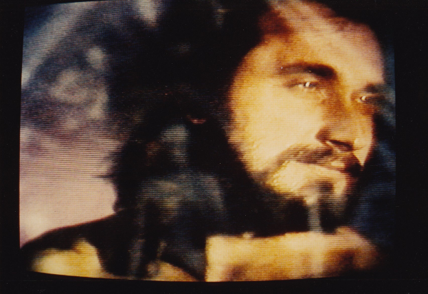 Ein Mann mit langem dunklem Bart und Haar, der ein weißes Hemd trägt, erscheint auf einem Fernseher mit dunklem Hintergrund und schaut direkt in die Kamera mit einem ernsten Gesichtsausdruck.