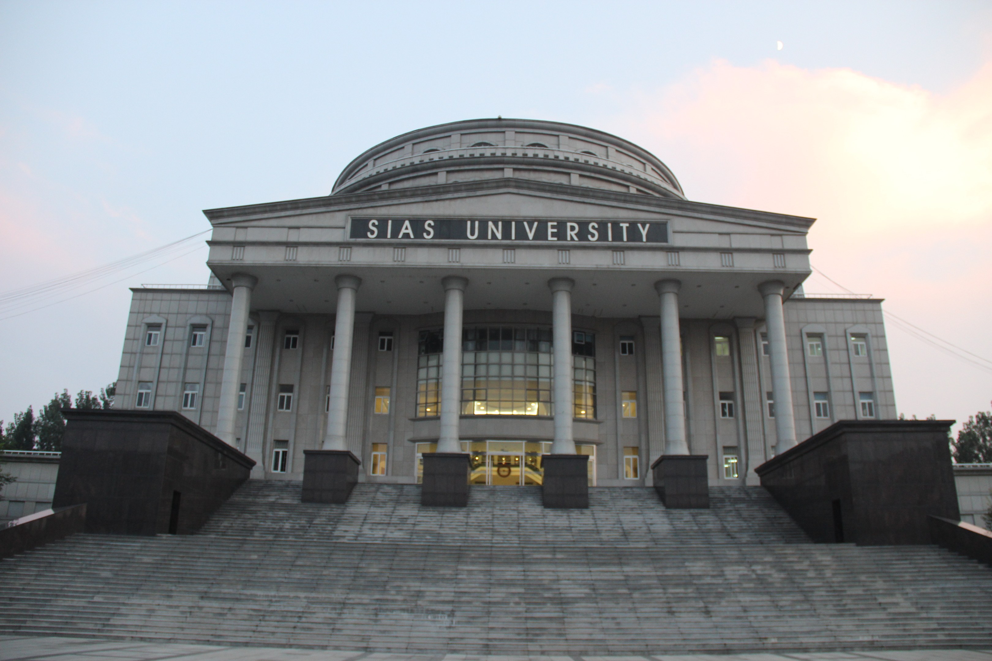 Großes Gebäude mit Säulen, Fenstern, Türen und Treppen, umgeben von Bäumen unter einem klaren blauen Himmel, mit einem Namen auf der Fassade, der wahrscheinlich eine Universität identifiziert.