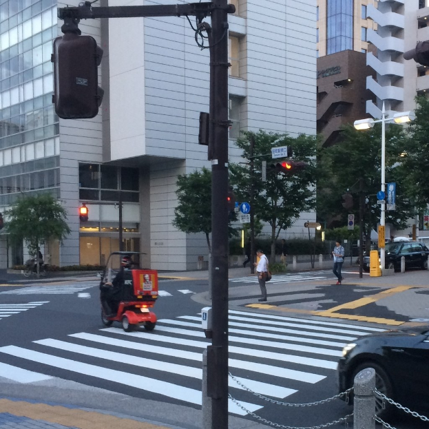 Ein E-Scooter auf einer Straße mit Personen im Hintergrund, einer Ampel im Vordergrund und Gebäuden mit Bäumen dahinter.