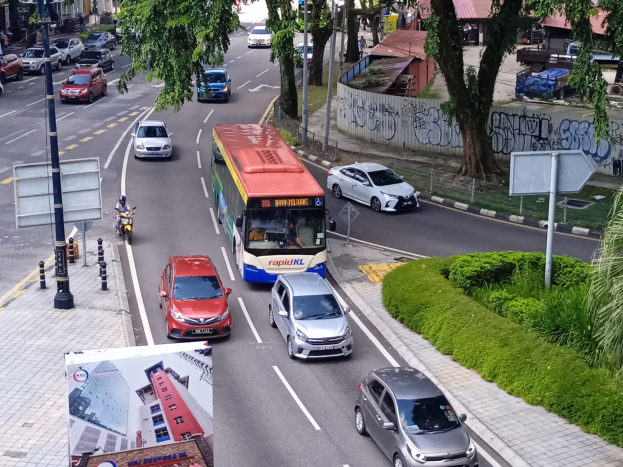 Eine belebte Stadtstraße mit Autos, Bussen, Motorrädern und Fußgängern, umgeben von Gebäuden, Bäumen und Straßeninfrastruktur.