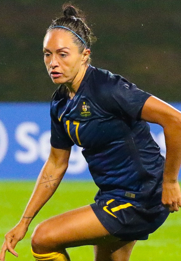 Eine Fußballspielerin in Aktion auf dem Feld, die ein blaues T-Shirt und gelbe Shorts trägt, mit einer blauen Tafel mit Text im Hintergrund.