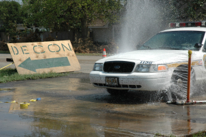 Ein Polizeiauto fährt durch eine Pfütze, mit einer Tafel, einem Verkehrskegel und anderen Gegenständen am Boden, mit Bäumen, Gebäuden und einem klaren blauen Himmel im Hintergrund.