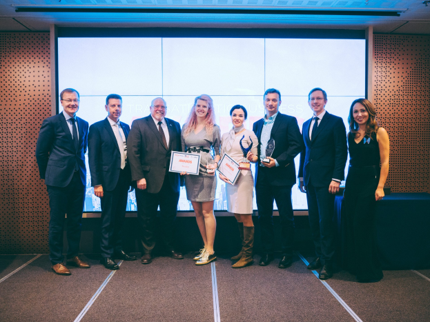 Gruppe von strahlenden Menschen mit Urkunden, die vor einem Bildschirm mit einem auf schwarzem Tuch gedeckten Tisch und einem Wandobjekt im Hintergrund stehen.