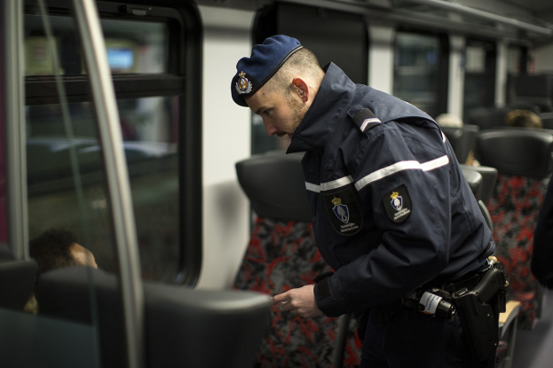 Polizeibeamter in Uniform und Mütze auf einem Zug stehend, der auf sein Handy schaut, mit sitzenden Passagieren und einer weiteren Person im Bild.