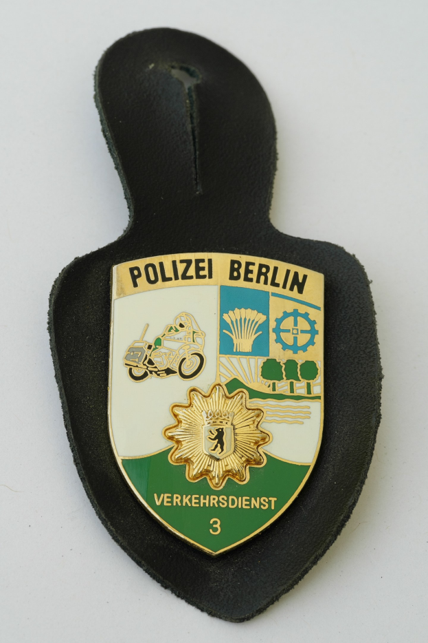 Ein schildförmiges Polizeisiegel mit "Polizei Berlin" in schwarzer fetter Schrift, das ein weißes Stern auf blauem Hintergrund und einen weißen Rand aufweist, das auf einer Oberfläche platziert ist.