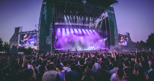 Eine große Menschenmenge versammelte sich auf einem Musikfestival, mit einer Bühne in der Mitte, auf der Musiker Instrumente spielen, beleuchtet von Lichtern und umgeben von Bildschirmen, mit einem Gebäude, Bäumen und einem klaren blauen Himmel im Hintergrund.