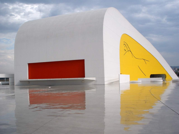 Das Museum of Contemporary Art in Mexiko-Stadt, ein großes weißes Gebäude mit einem gelben und roten Wandgemälde an der Seite, umgeben von Wasser mit Bäumen, Bergen und einem klaren blauen Himmel im Hintergrund.