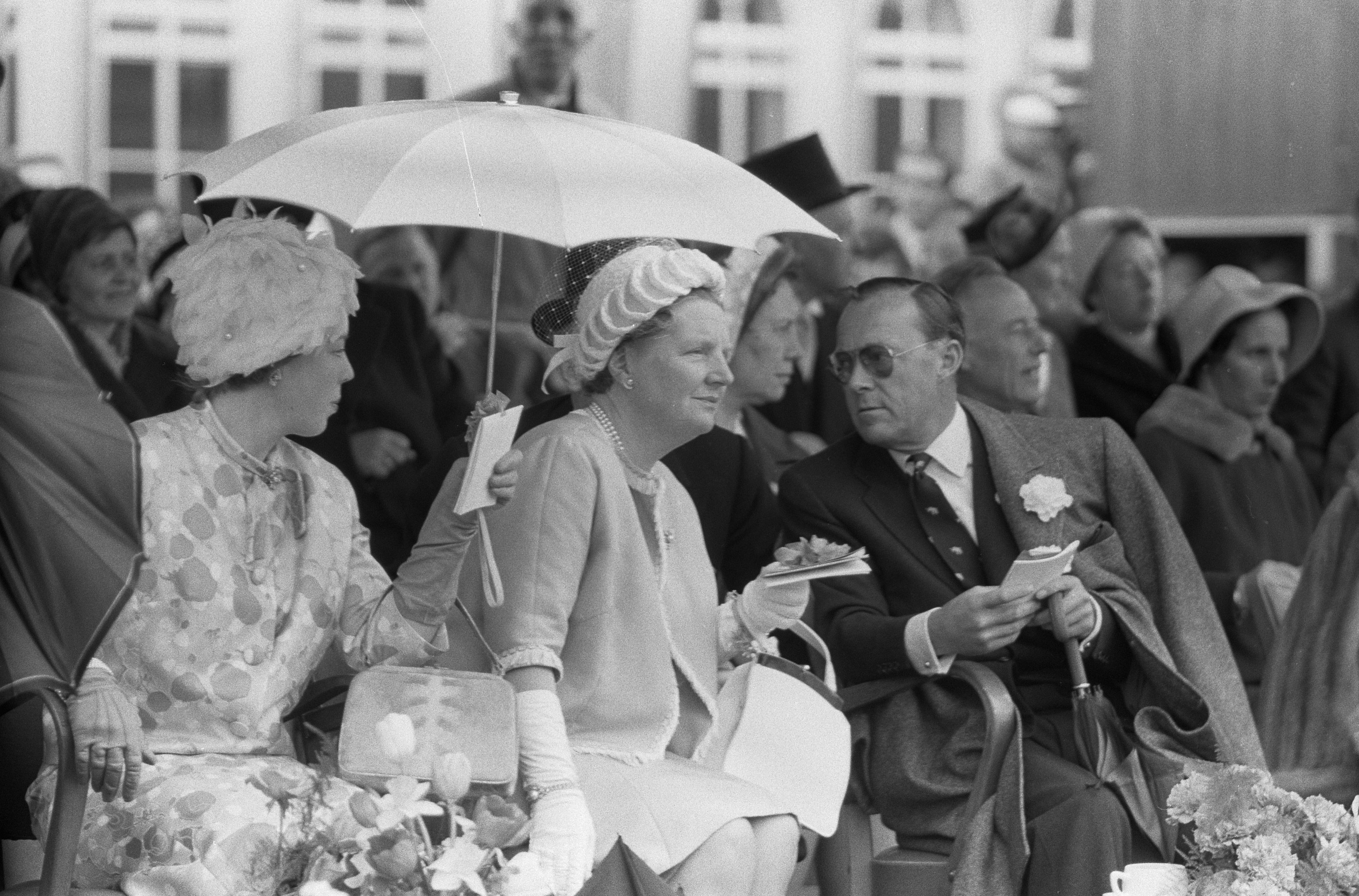 Schwarzes und weißes Foto von Queen Elizabeth II und Prinz Philip, die mit Schirmen auf Stühlen sitzen, umgeben von einer Menge mit Hüten und Büchern, mit einem Gebäude und Blumenschmuck im Hintergrund.