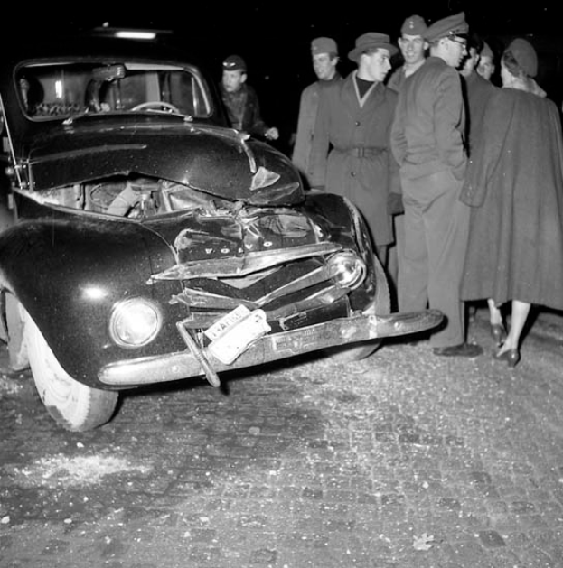 Eine Schwarz-Weiß-Szene eines beschädigten Autos am Straßenrand, umgeben von einer Gruppe besorgter Menschen.