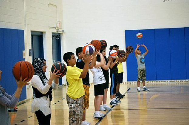 Gruppe junger Menschen auf einem Basketballfeld mit Basketballs, im Hintergrund Türen und eine Wand.