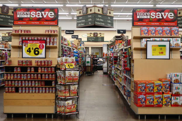 Ein Innenraum eines Lebensmittelladens mit Regalen und Boden voller verschiedener Lebensmittel, Deckenbeleuchtung und einer nummerierten Tafel im Hintergrund.