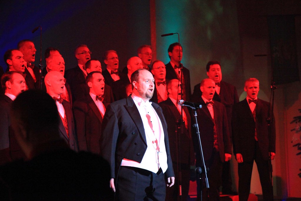Eine Gruppe von Menschen, die vor Ständern und Mikrofonen singen, mit einer Wand im Hintergrund.