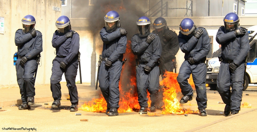 Menschen in Helmen stehen vor einem Feuer, mit Gebäuden, einem Fahrzeug und Gegenständen auf der Oberfläche dahinter; ein Plakat und eine Tafel sind an der linken Wand, und Text steht unten auf dem Bild.