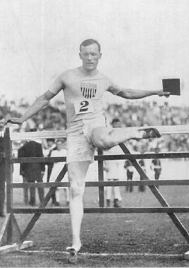 Schwarz-weiß-Foto eines Mannes in Sportkleidung, der über ein Hindernis springt, mit einer Nummer auf seinem Hemd und Shorts, vor Zuschauern unter freiem Himmel.