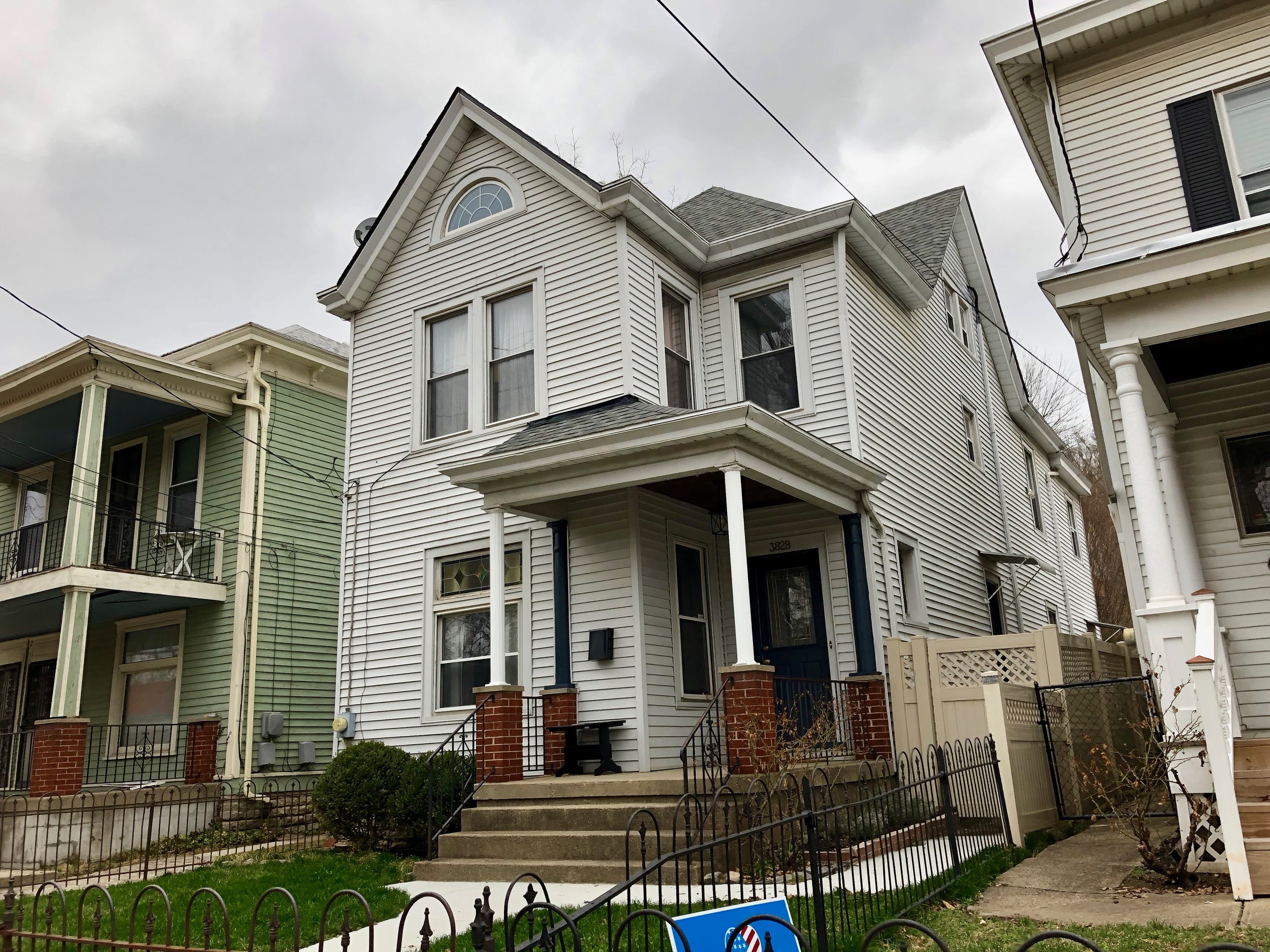 Reihe von Häusern mit Fenstern, Türen, Säulen, Geländern, Treppen und Dächern, umgeben von einem Metallzaun, Gras, Pflanzen, Bäumen und einem bewölkten Himmel, mit einem Mietschild unten, das die Adresse und Details auflistet.