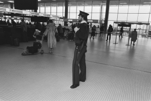 Polizeibeamter in Uniform mit einer Waffe in der Hand, der in einem Flughafen-Terminal steht, umgeben von Menschen und Gepäck, mit Gepäckwagen, Deckenleuchten, Pfosten, Informationsanzeigen und Glaswänden im Hintergrund.
