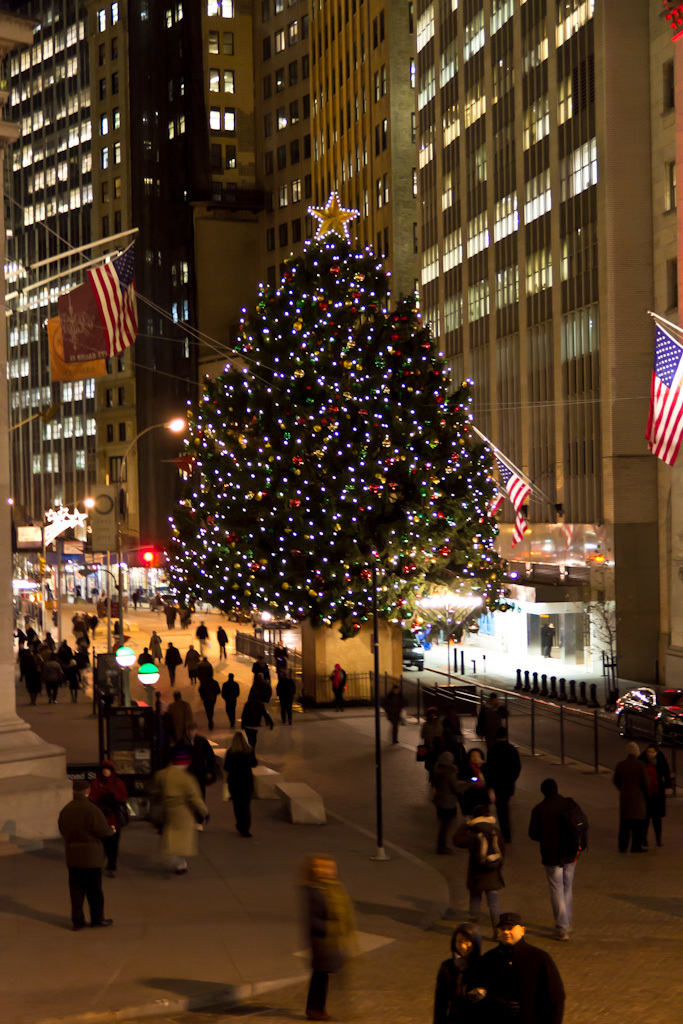 Ein mit Menschen geschmückter Weihnachtsbaum steht in der Mitte einer Straße, flankiert von zwei Fahnenmasten auf beiden Seiten, mit beleuchteten Gebäuden im Hintergrund.