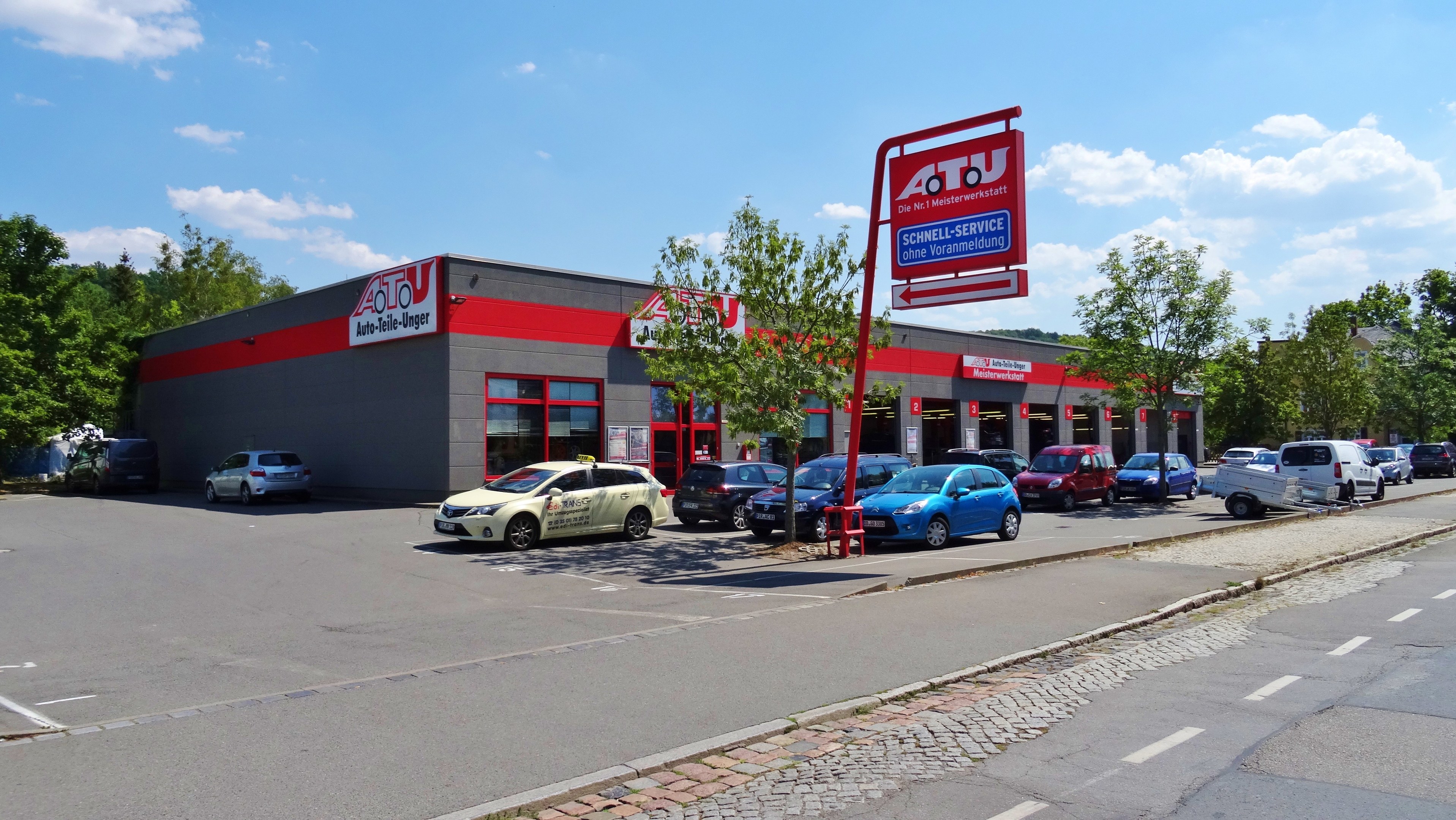 Ein Autohaus mit mehreren geparkten Autos vor einem Gebäude, umgeben von weiteren Gebäuden, Bäumen, Strommasten, Schildern und einer Straße unter einem bewölkten Himmel.